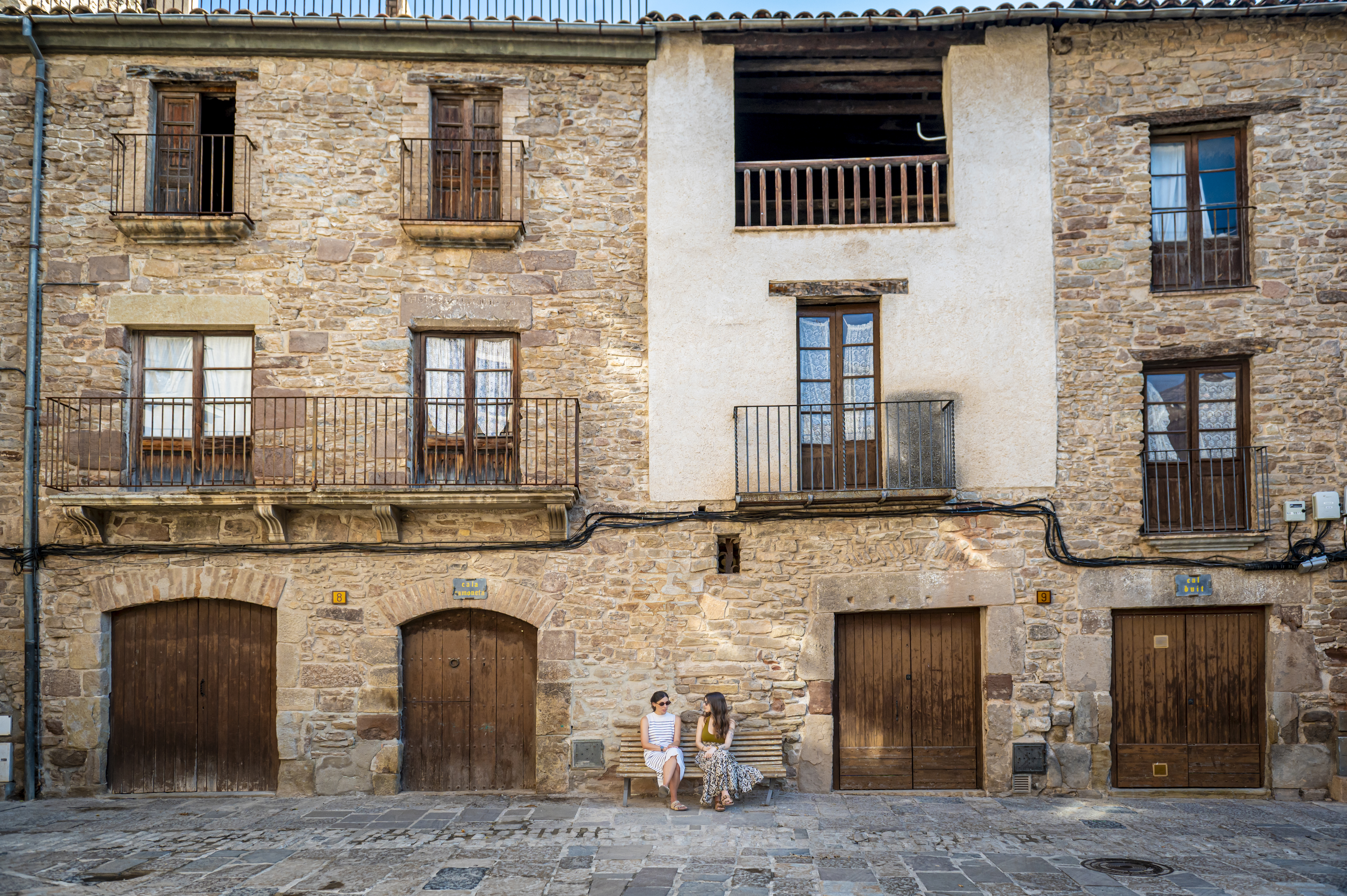 Edificis empedrats de la Plaça Major del Poble Vell amb un banc i dos noies assegudes en ell.